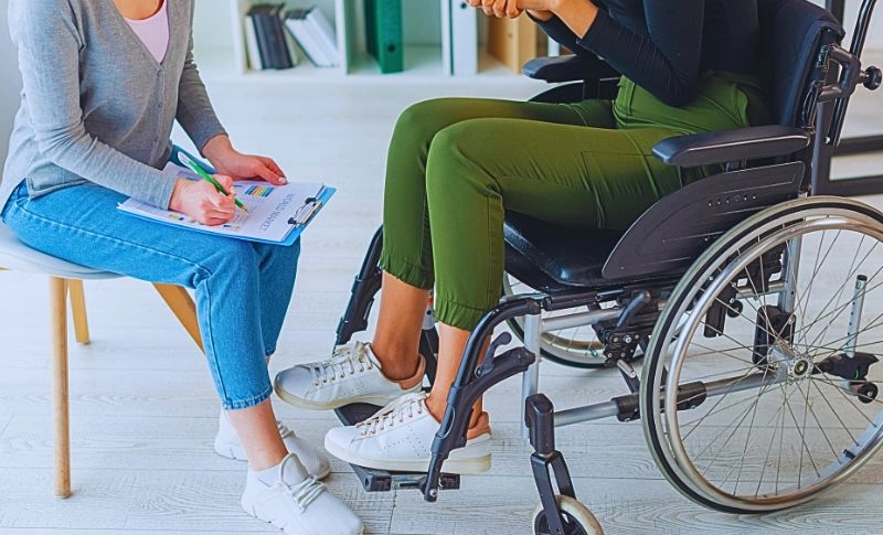 Woman in wheelchair talking to another woman holding a pen and a clipboard.