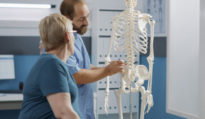 Helping Patients Understand the Connection Between Injuries and Long-Term Health 1 Medical professional pointing at a human skeleton model, specifically highlighting the spinal cord area.