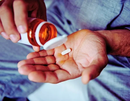a person pouring a single white capsule pill from an amber prescription bottle into their open palm.